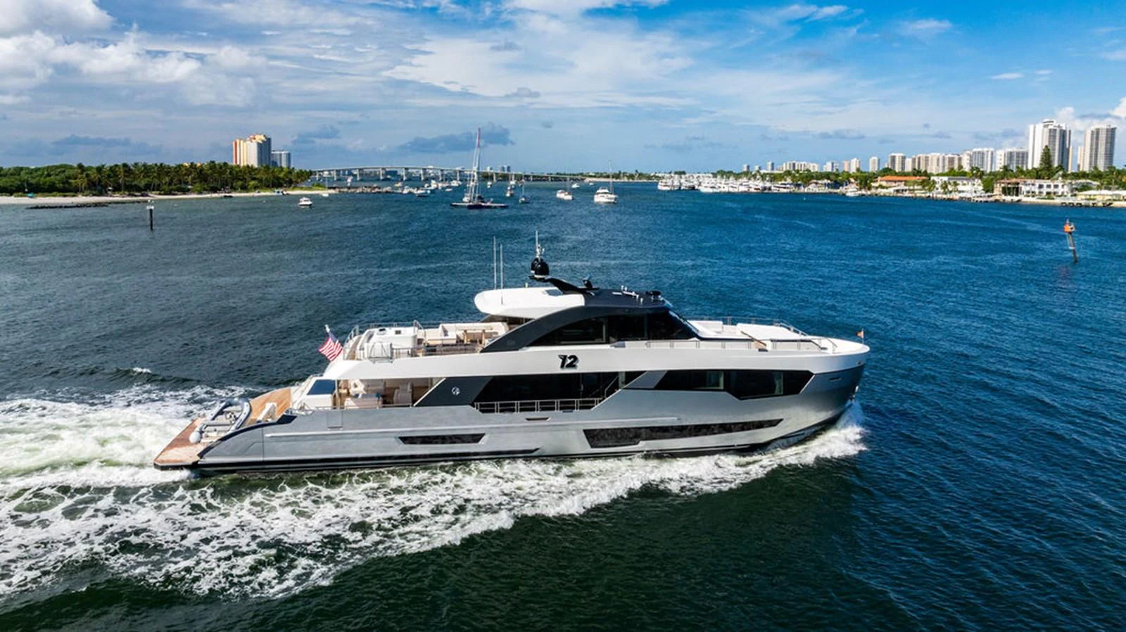 A sleek and modern yacht cruises through a scenic coastal area with a city skyline in the background. aboard M/Y 12 Yacht for Sale