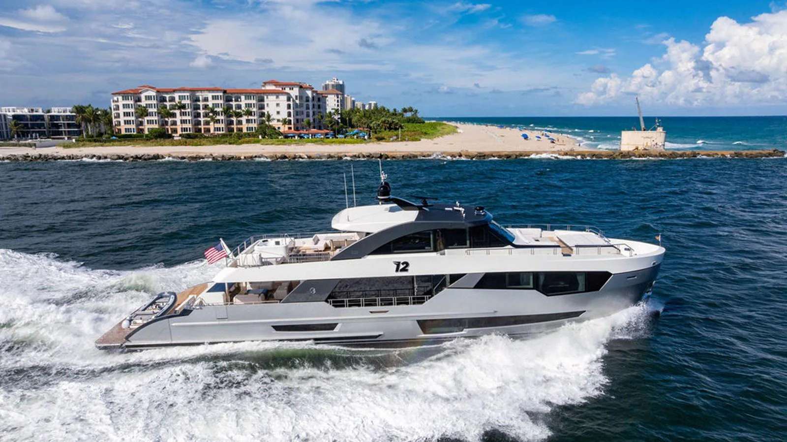 A sleek and modern yacht cruises along the coastline with a backdrop of a beach and residential buildings. aboard M/Y 12 Yacht for Sale