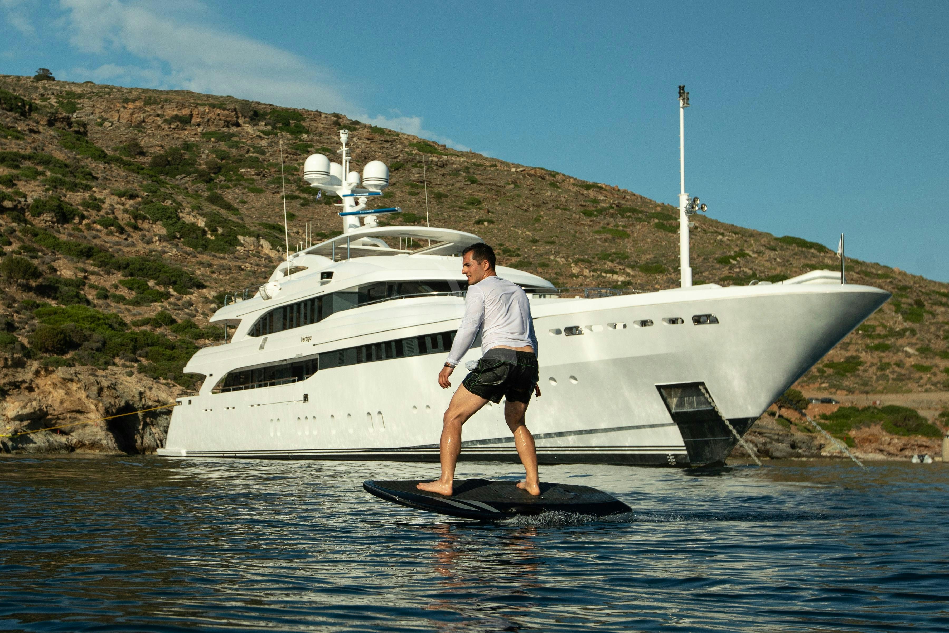 a man standing on a surfboard in front of a large white ship aboard VERTIGO Yacht for Charter