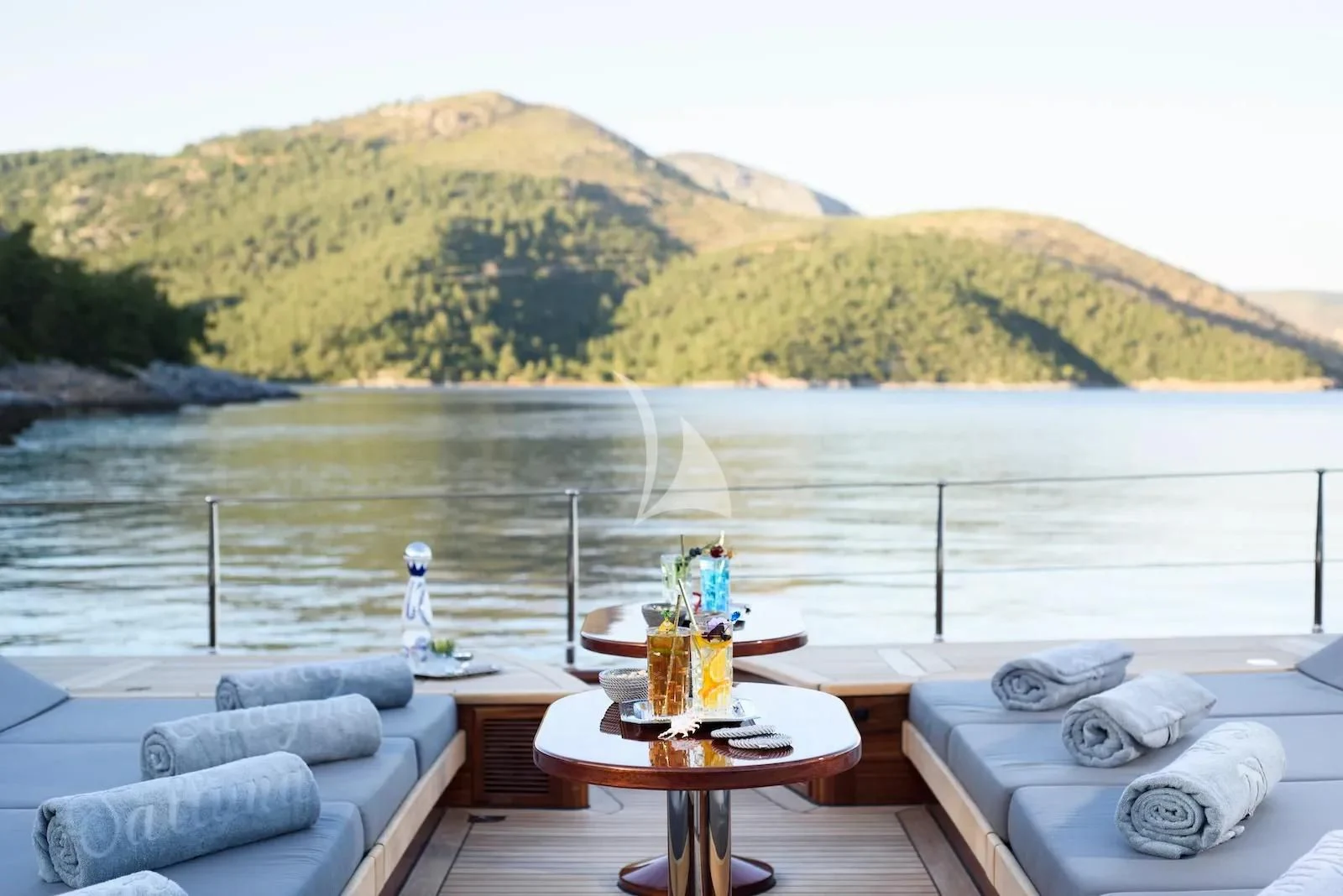 a table with a glass of beer on a beach with a body of water and hills in the aboard SALLYNA Yacht for Sale