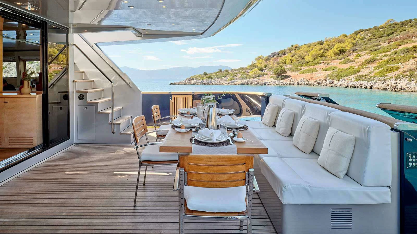 a large white table with chairs on a deck with a view of a mountain range aboard AMICI PER SEMPRE Yacht for Charter