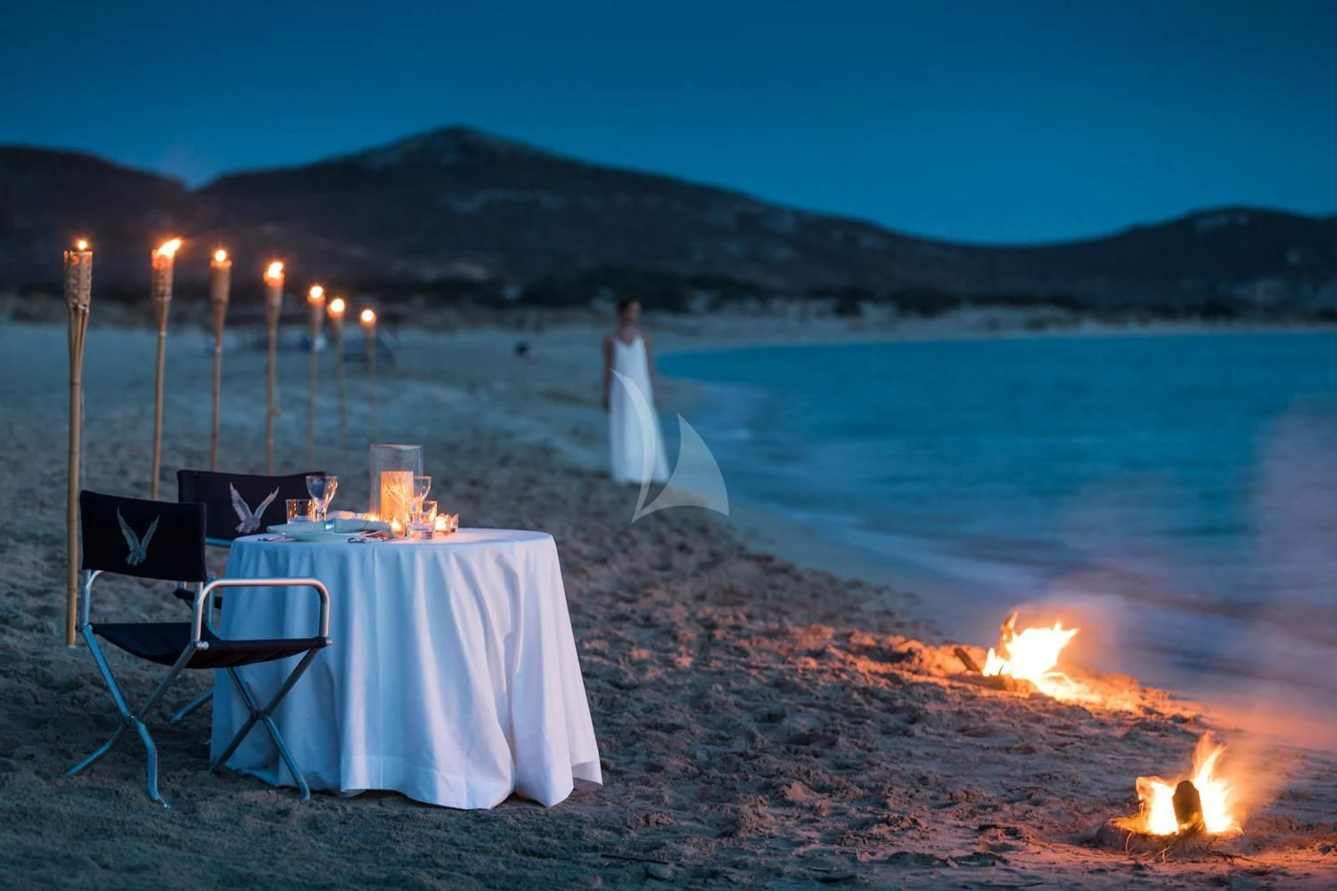 a table set up with candles on a beach with a body of water and mountains in the background aboard MALTESE FALCON Yacht for Sale