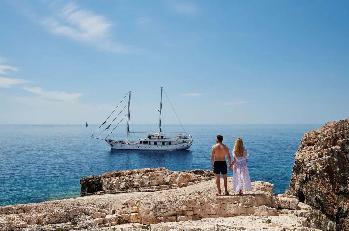 a man and woman walking on a rocky beach with a boat in the water aboard CORSARIO Yacht for Charter