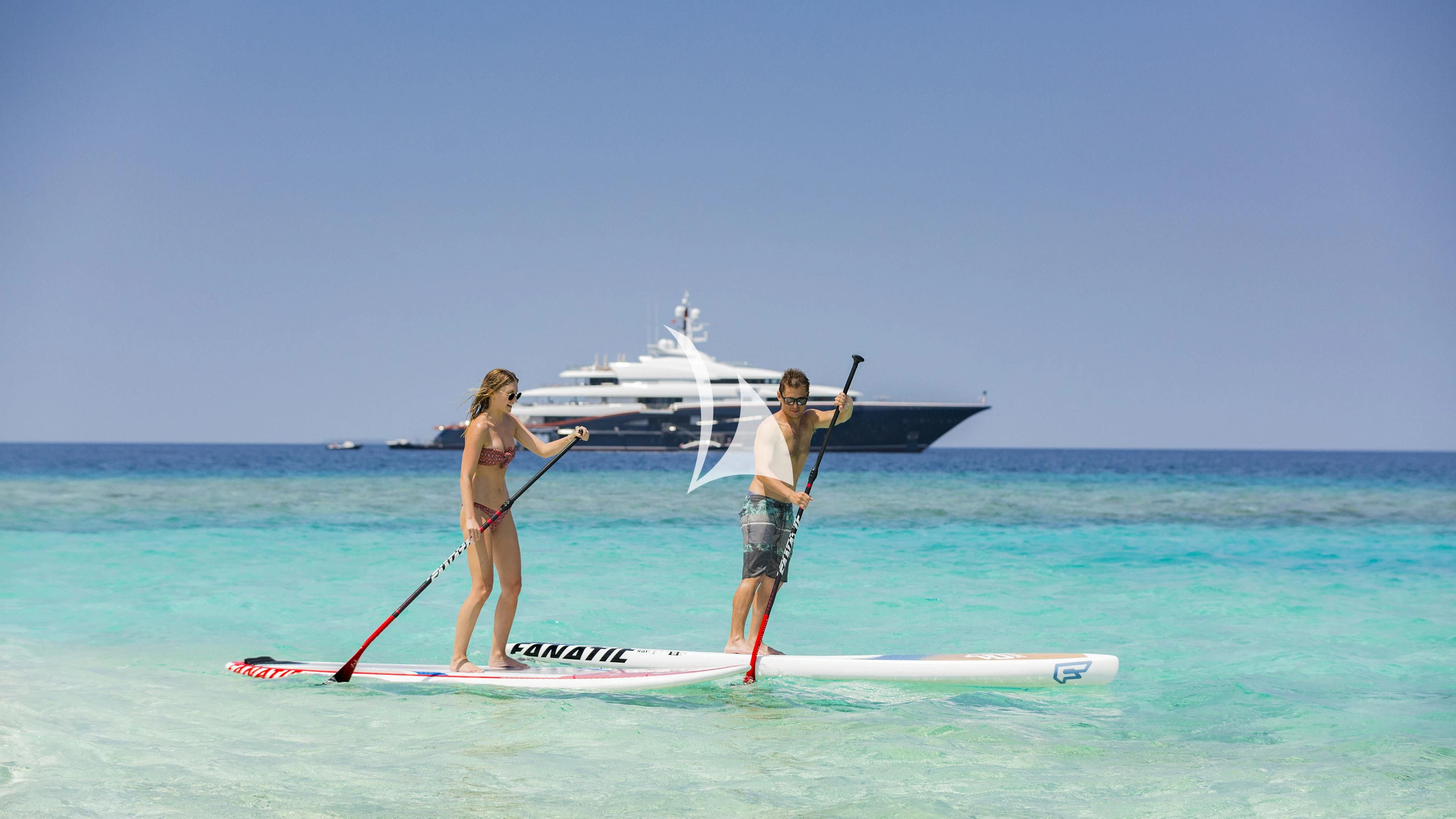 a man and woman on a surfboard with a sailboat in the background aboard NIRVANA Yacht for Sale
