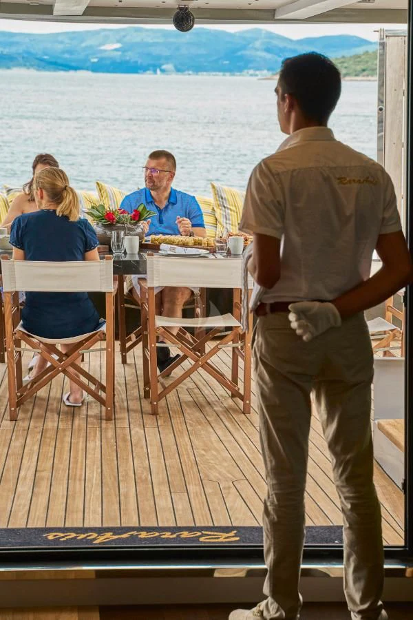 a man standing on a deck looking at a woman sitting at a table aboard RARA AVIS Yacht for Charter