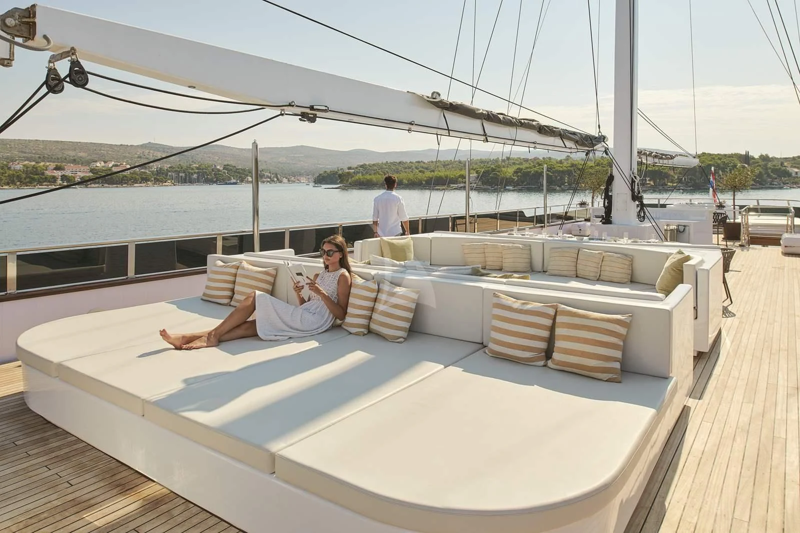 a woman sitting on a lounge chair on a deck with a large white sailboat and a man aboard ANIMA MARIS Yacht for Sale
