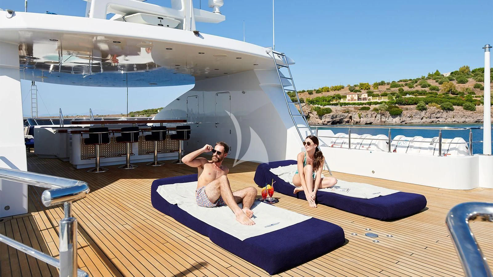 a man and woman sitting on a mat on a deck with a boat in the background aboard RARE FIND Yacht for Charter