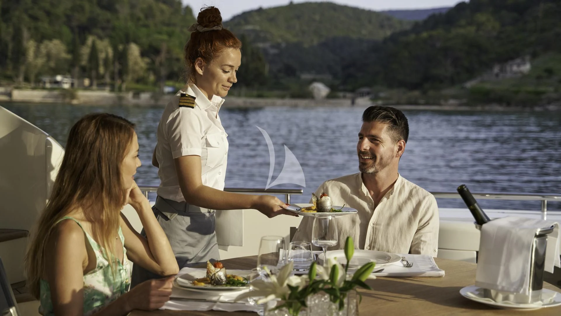 a man and woman sitting at a table with a plate of food and water and mountains in the aboard ACCEPTUS Yacht for Sale