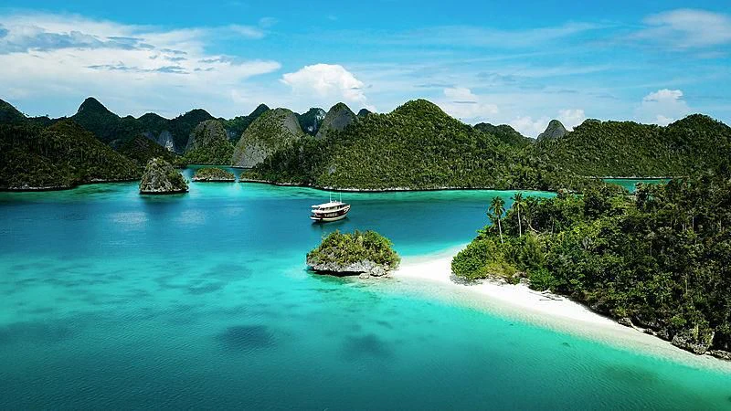 a blue lagoon with a boat and islands with Raja Ampat Islands in the background aboard MISCHIEF Yacht for Sale