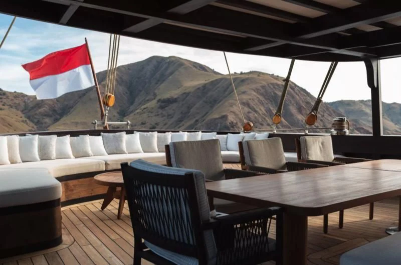 a deck with a table and chairs and a mountain in the background aboard VELA Yacht for Charter