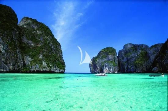 a body of water with a rocky cliff and a boat in it with Phi Phi Islands in the background aboard MIA KAI Yacht for Charter