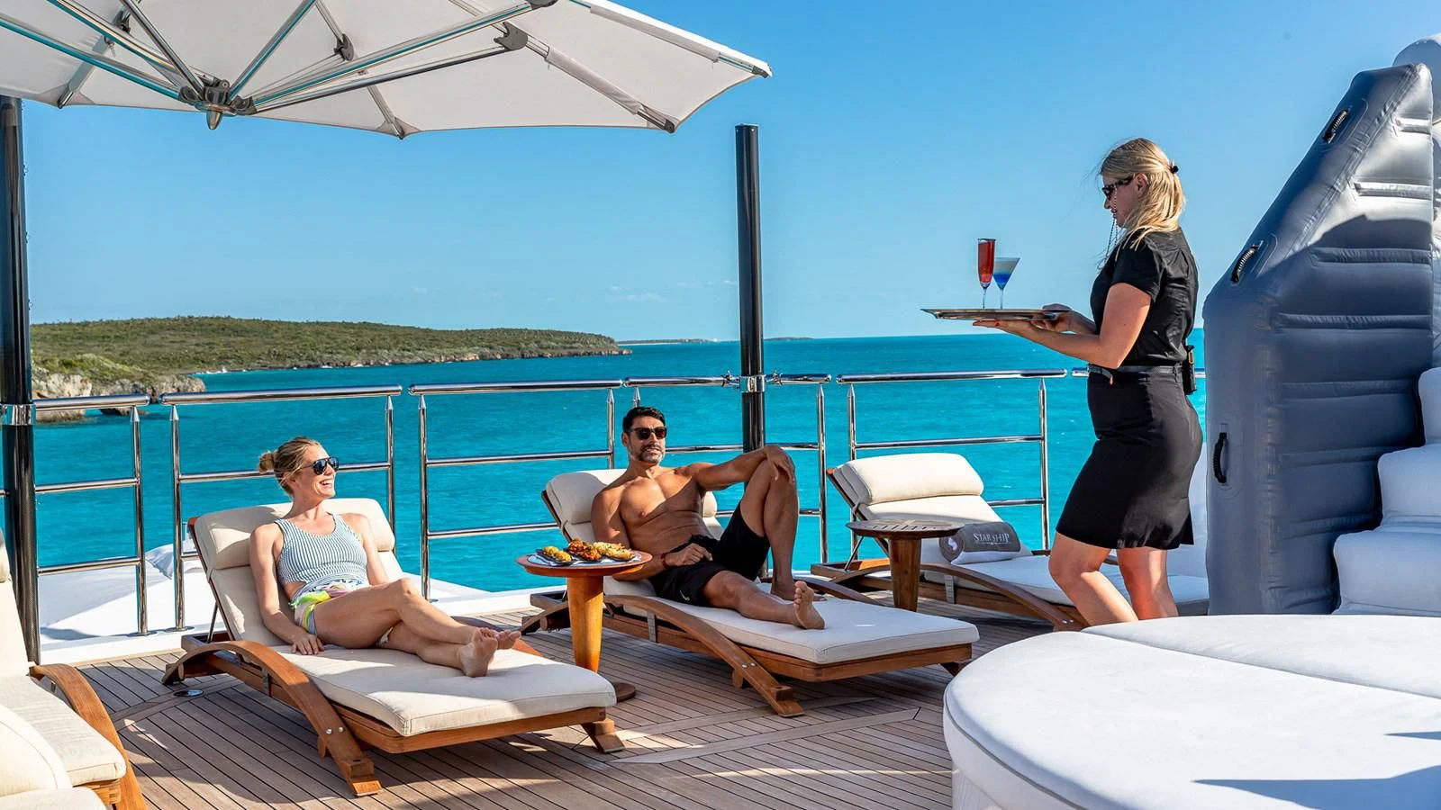 a group of people on a lounge chair on a deck with a body of water and a beach aboard STARSHIP Yacht for Charter