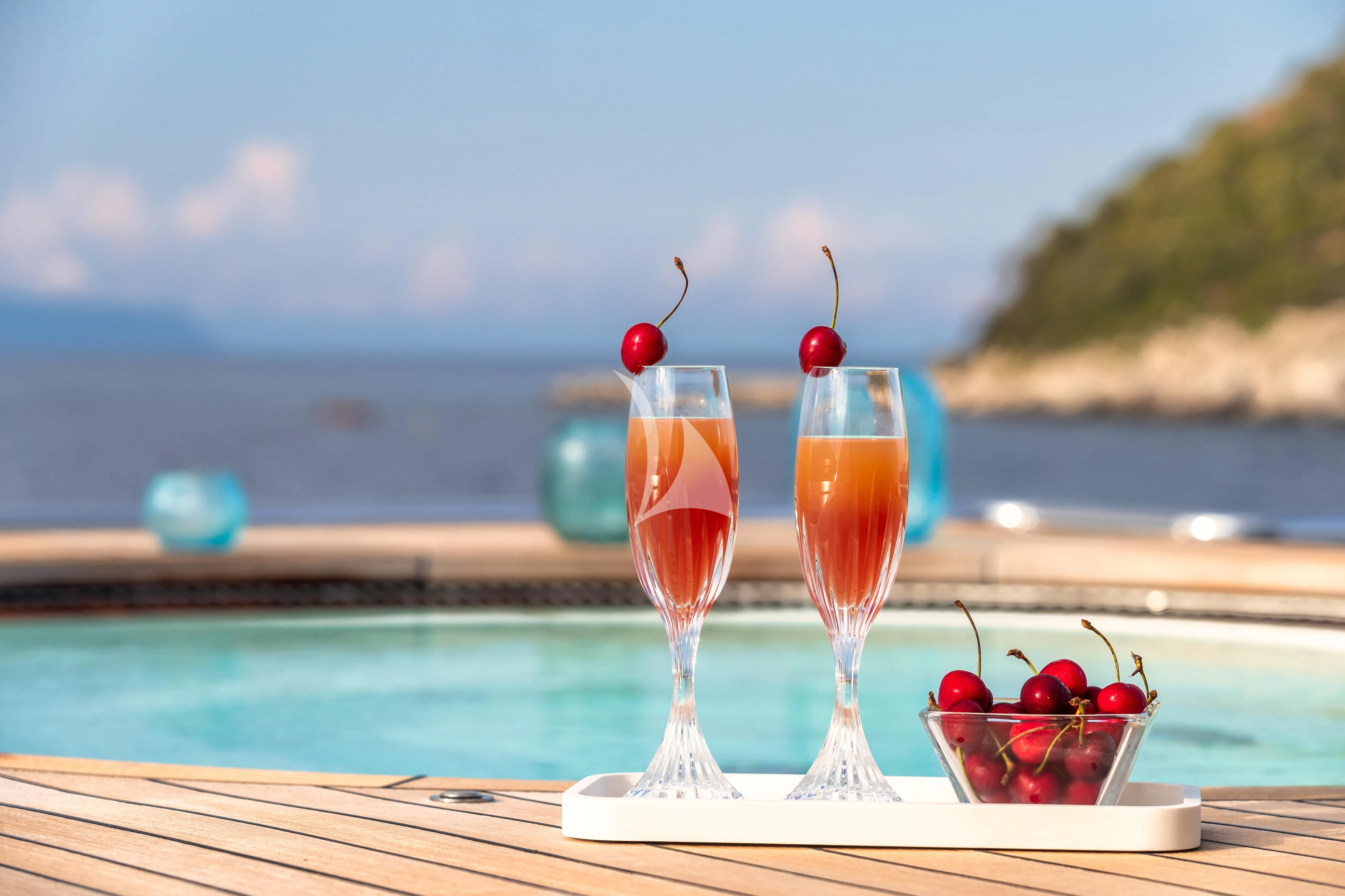 a couple of glasses with red liquid and fruit on a table by a body of water aboard PEARL Yacht for Sale