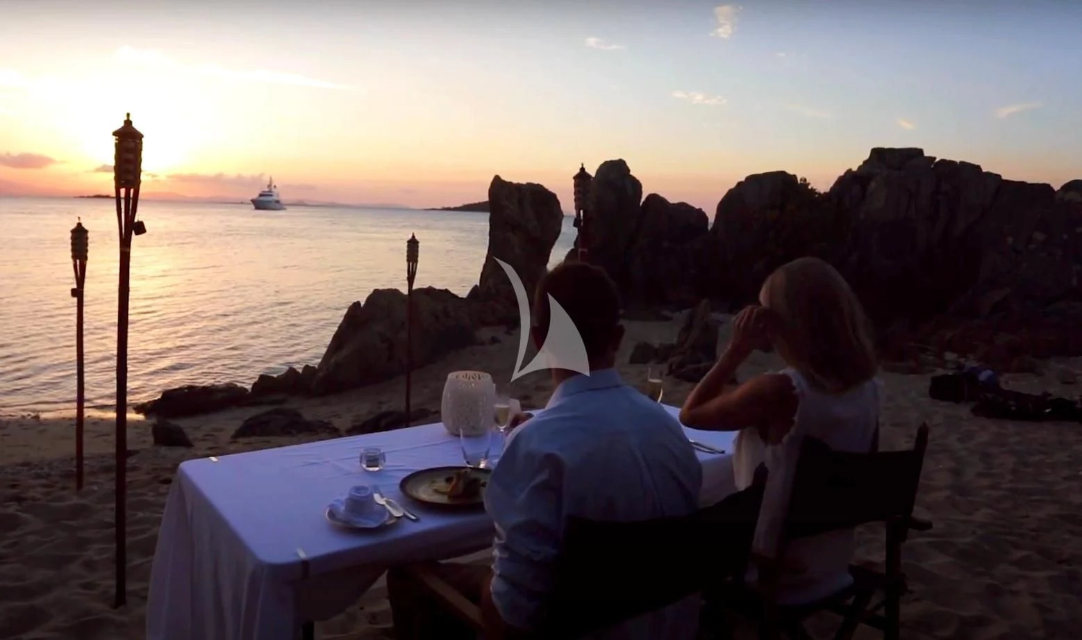 a man and woman sitting at a table with a large rock formation in the background aboard DE LISLE III Yacht for Sale