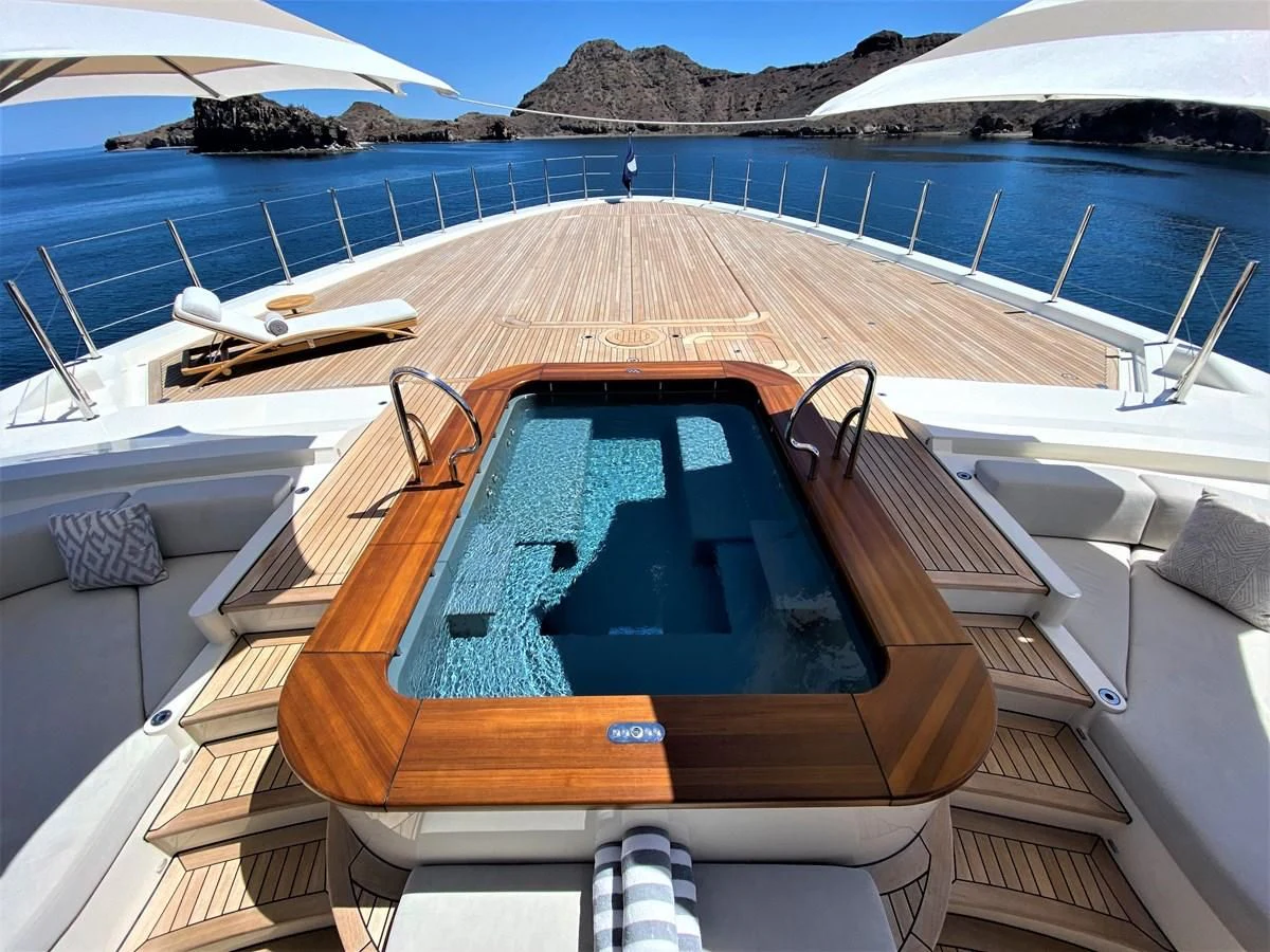 a swimming pool with a deck and chairs and a beach and mountains in the background aboard MAN OF STEEL Yacht for Charter