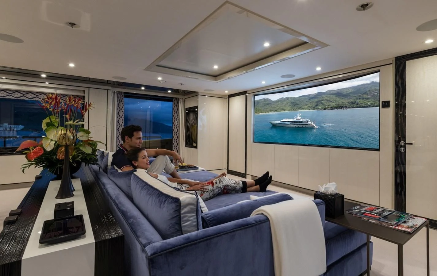 a man and a woman sitting on a couch in a room with a large window and a large aboard AMIGOS Yacht for Charter