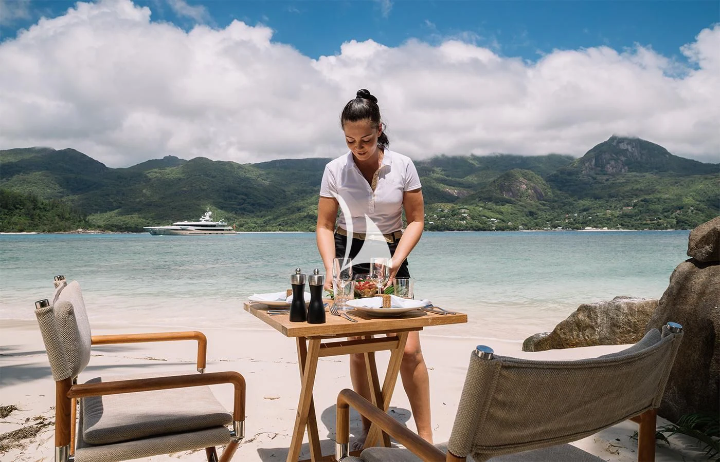 a person standing at a table with wine bottles and a glass of wine on a beach with a aboard AMIGOS Yacht for Charter