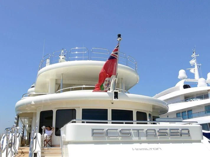 a person sitting on a chair on a boat with a flag on the front aboard ECSTASEA Yacht for Sale