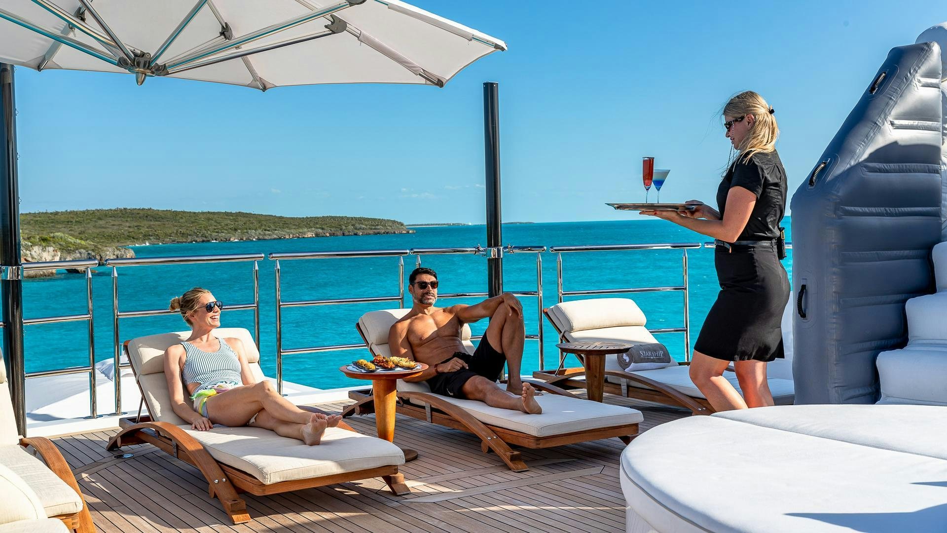 a group of people on a lounge chair on a deck with a body of water and a beach aboard STARSHIP Yacht for Sale