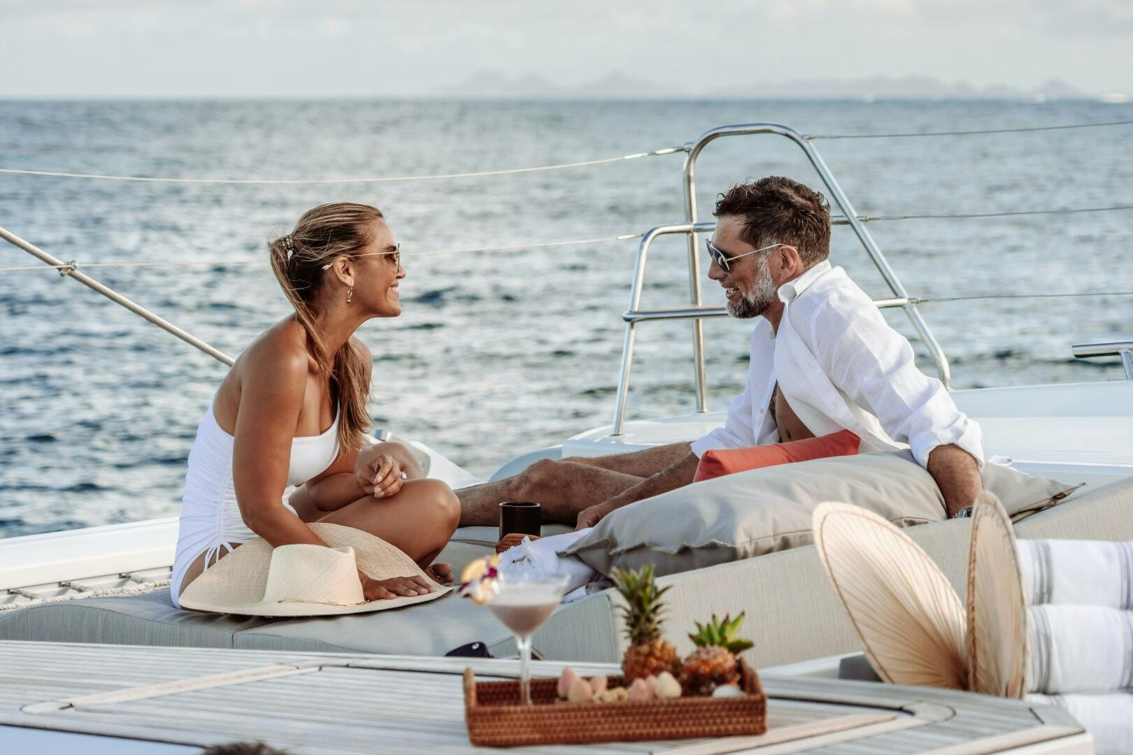 a man and woman sitting on a boat with a drink and a picnic table on the beach aboard OCEANUS Yacht for Sale