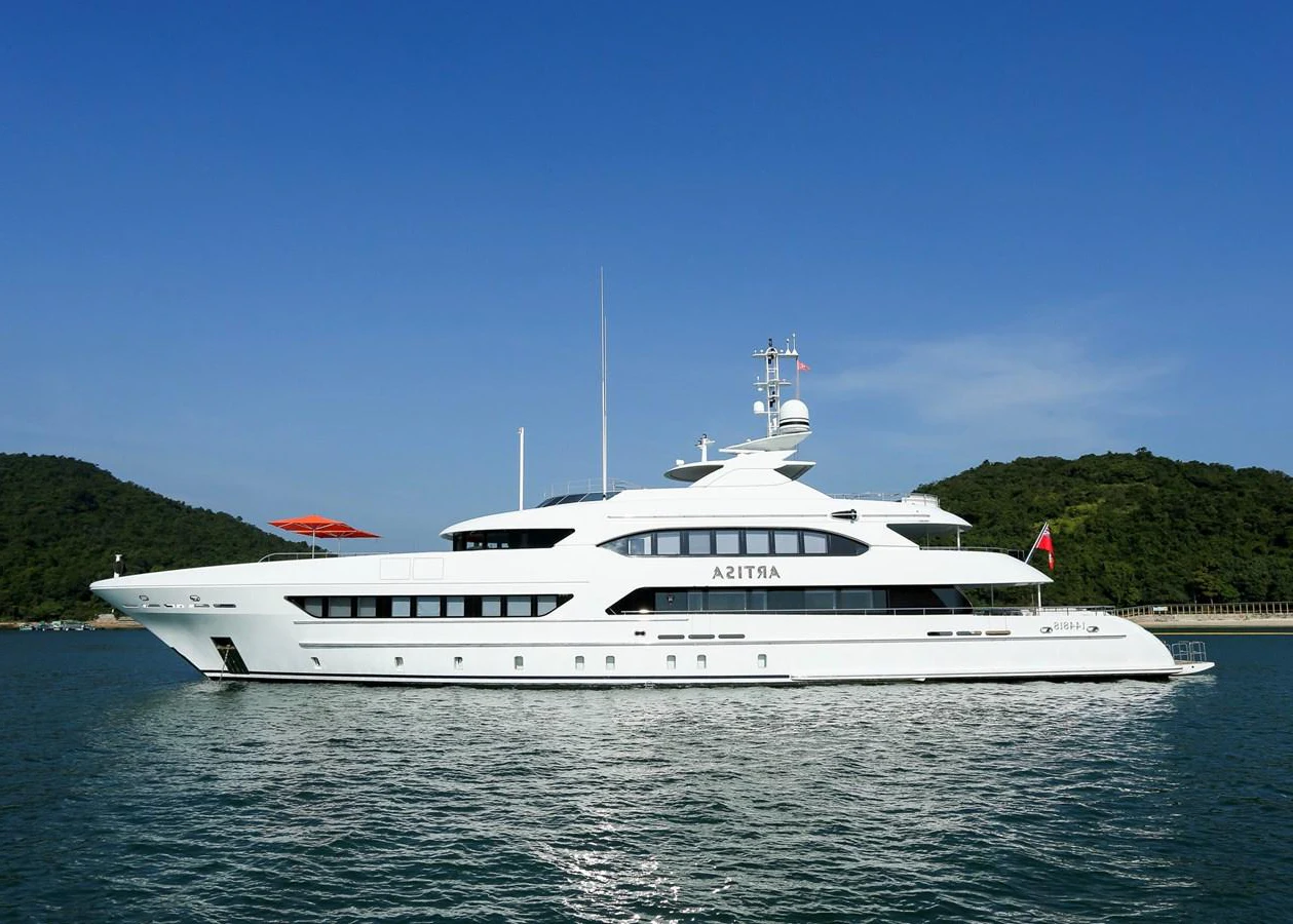 A sleek white yacht cruises gracefully on calm waters against a backdrop of lush green hills and a clear blue sky. aboard HEESEN 47M Yacht for Sale