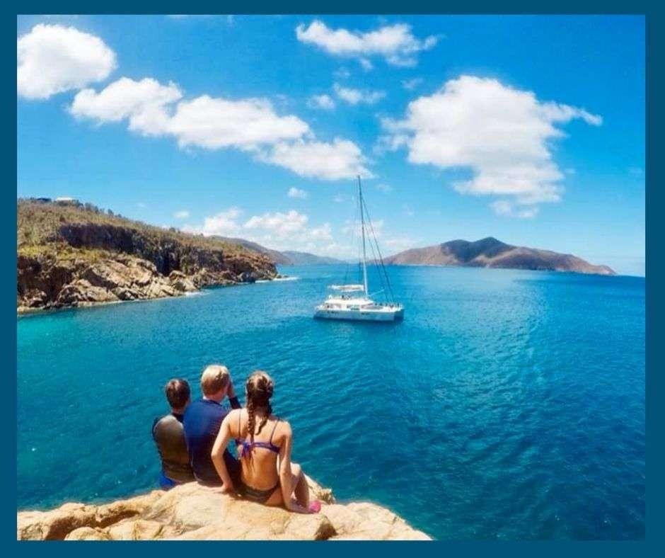a group of people sitting on a rock in the water with a sailboat in the background aboard VIRAMAR Yacht for Charter