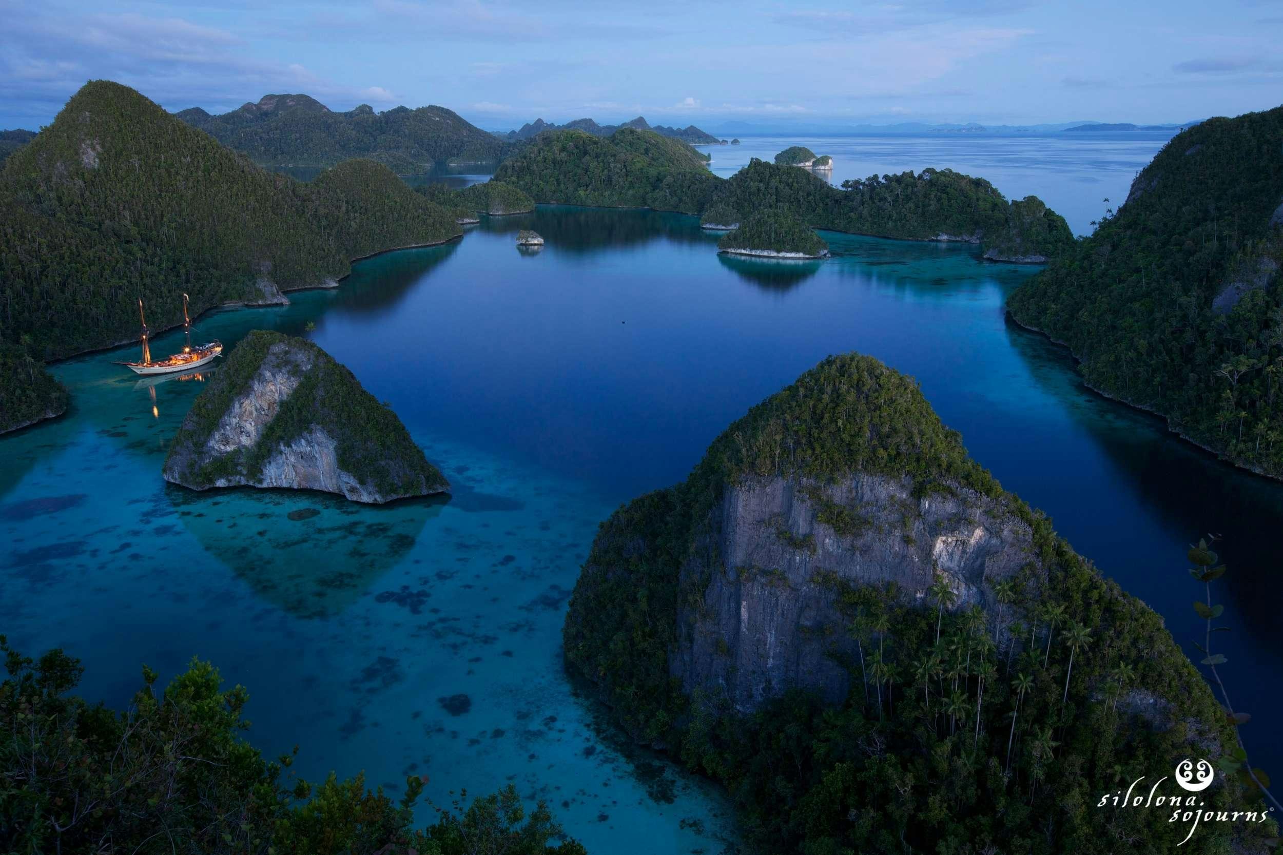 a body of water with islands and boats in it with Raja Ampat Islands in the background aboard SILOLONA Yacht for Charter