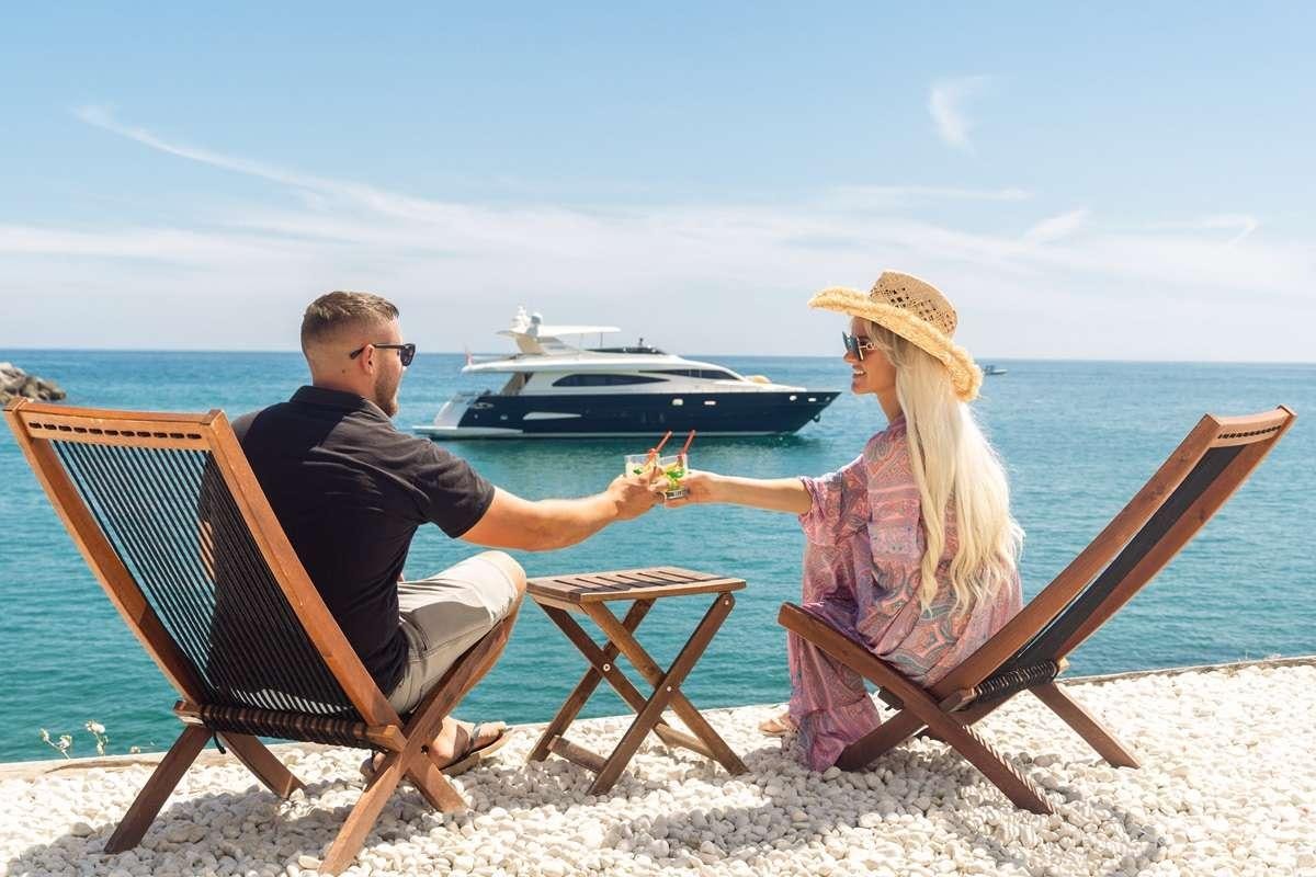 a man and woman sitting on a bench at the beach with a boat in the background aboard M Yacht for Charter