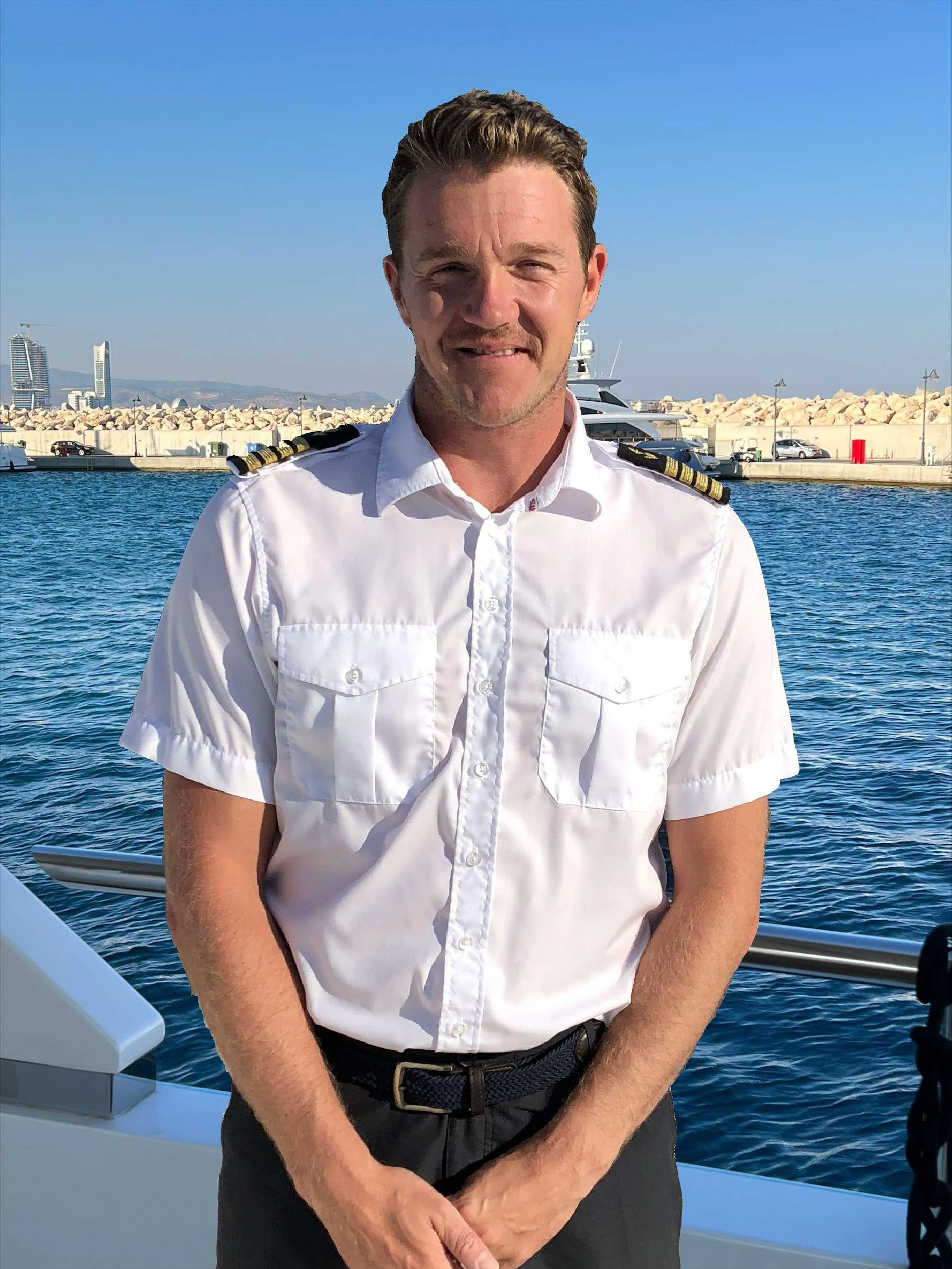 A crew member stands on the deck of a yacht with a marina in the background. aboard LE VERSEAU Yacht for Charter