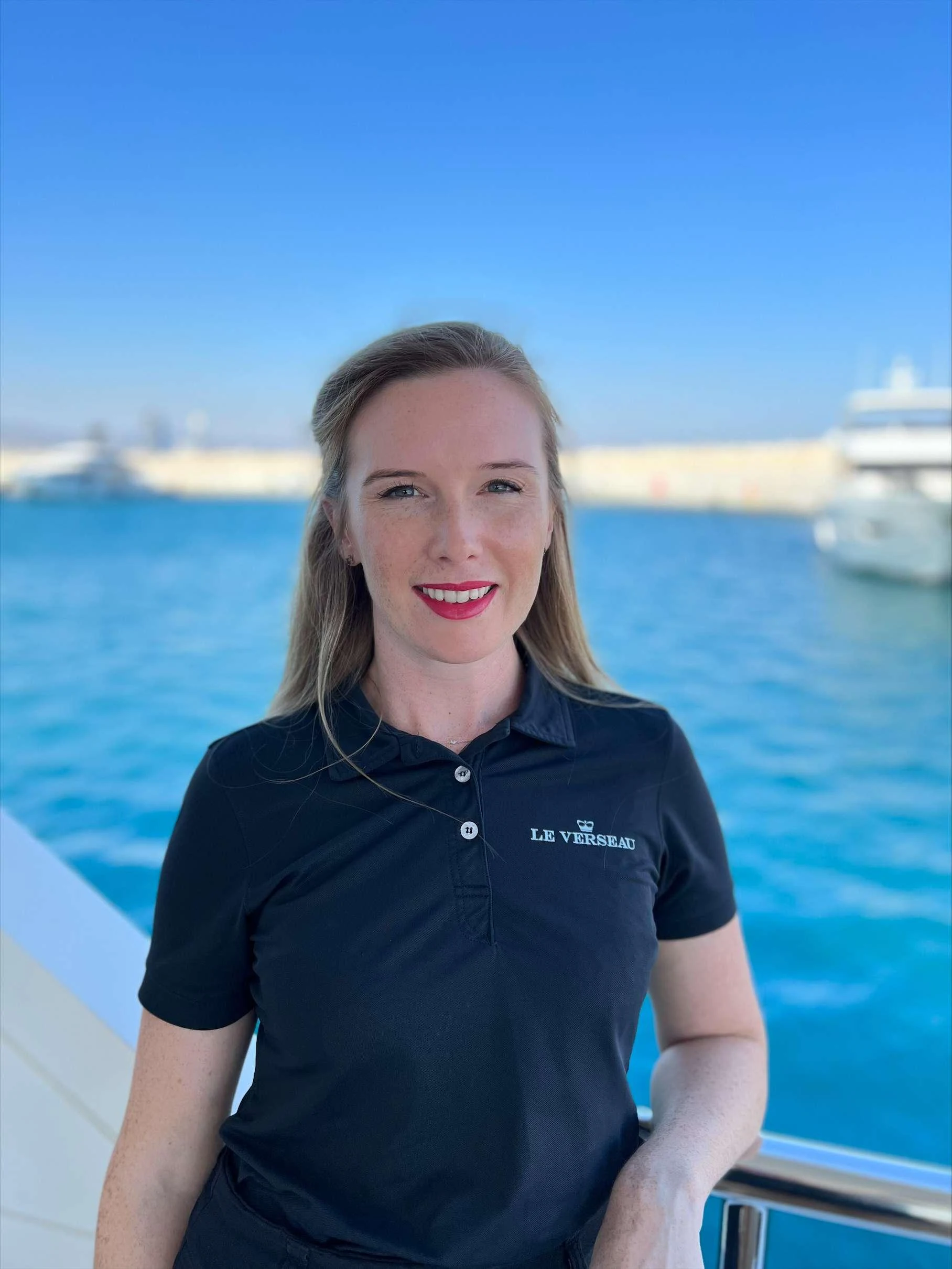 A crew member stands on the deck of the yacht with a marina in the background. aboard LE VERSEAU Yacht for Charter
