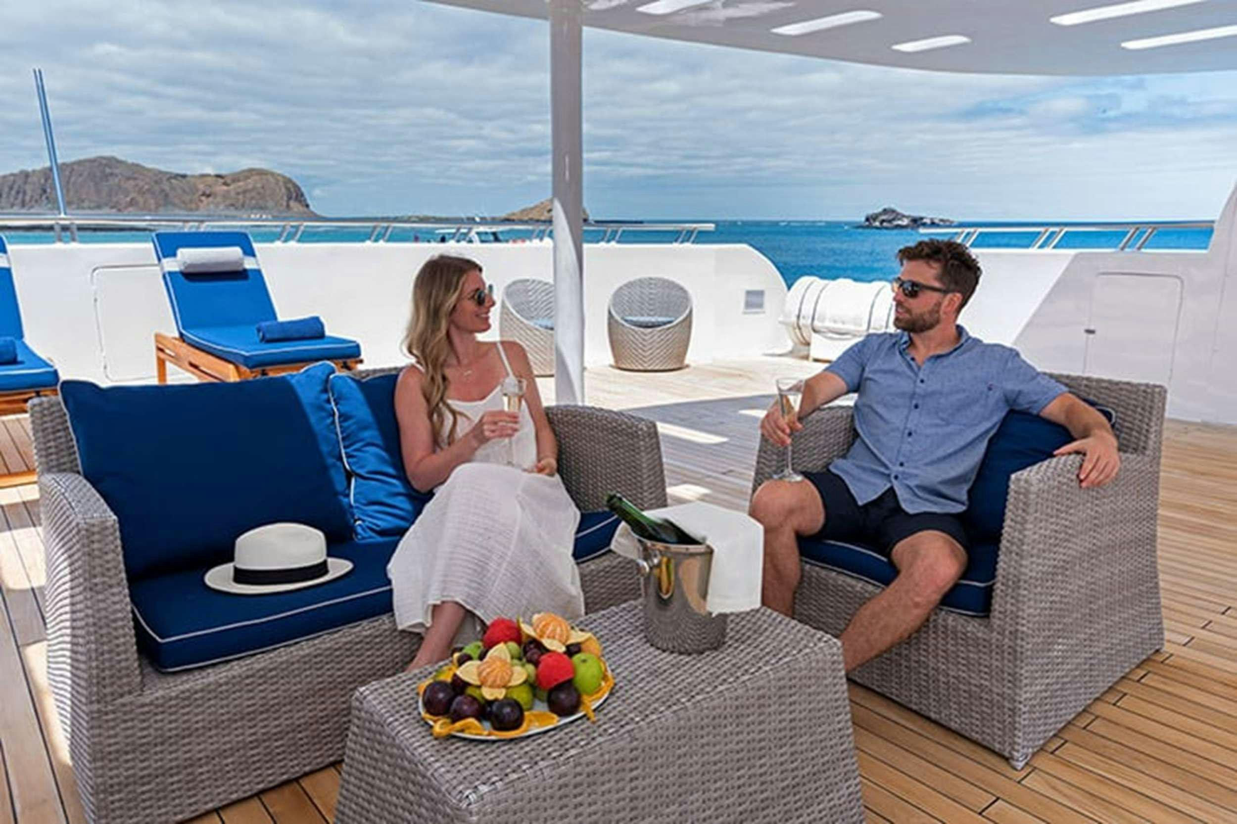 a man and woman sitting on a couch next to a table with fruit on it aboard GALAPAGOS HORIZON Yacht for Charter