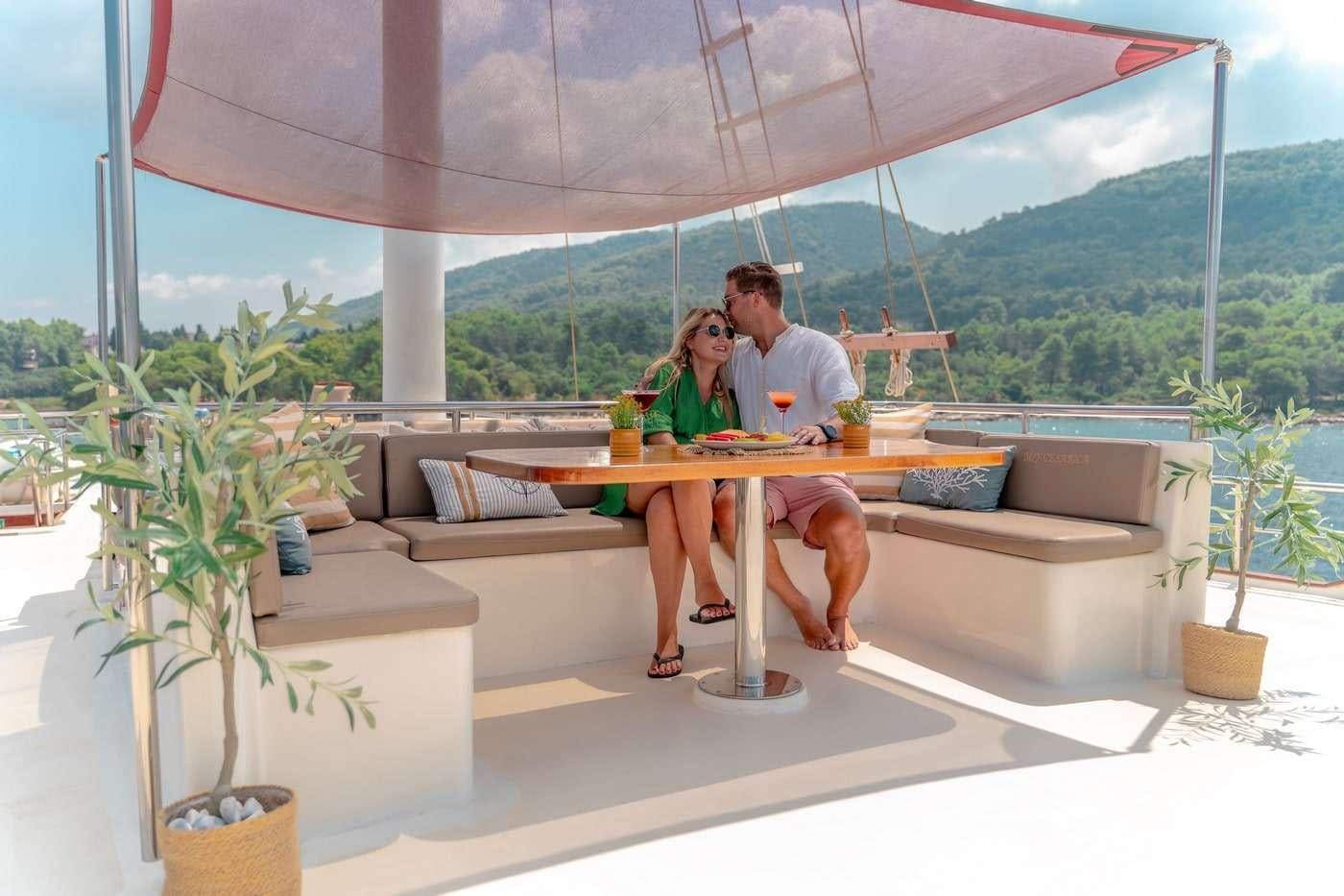 a man and a woman sitting on a couch in a room with a view of mountains and trees aboard CESARICA Yacht for Charter