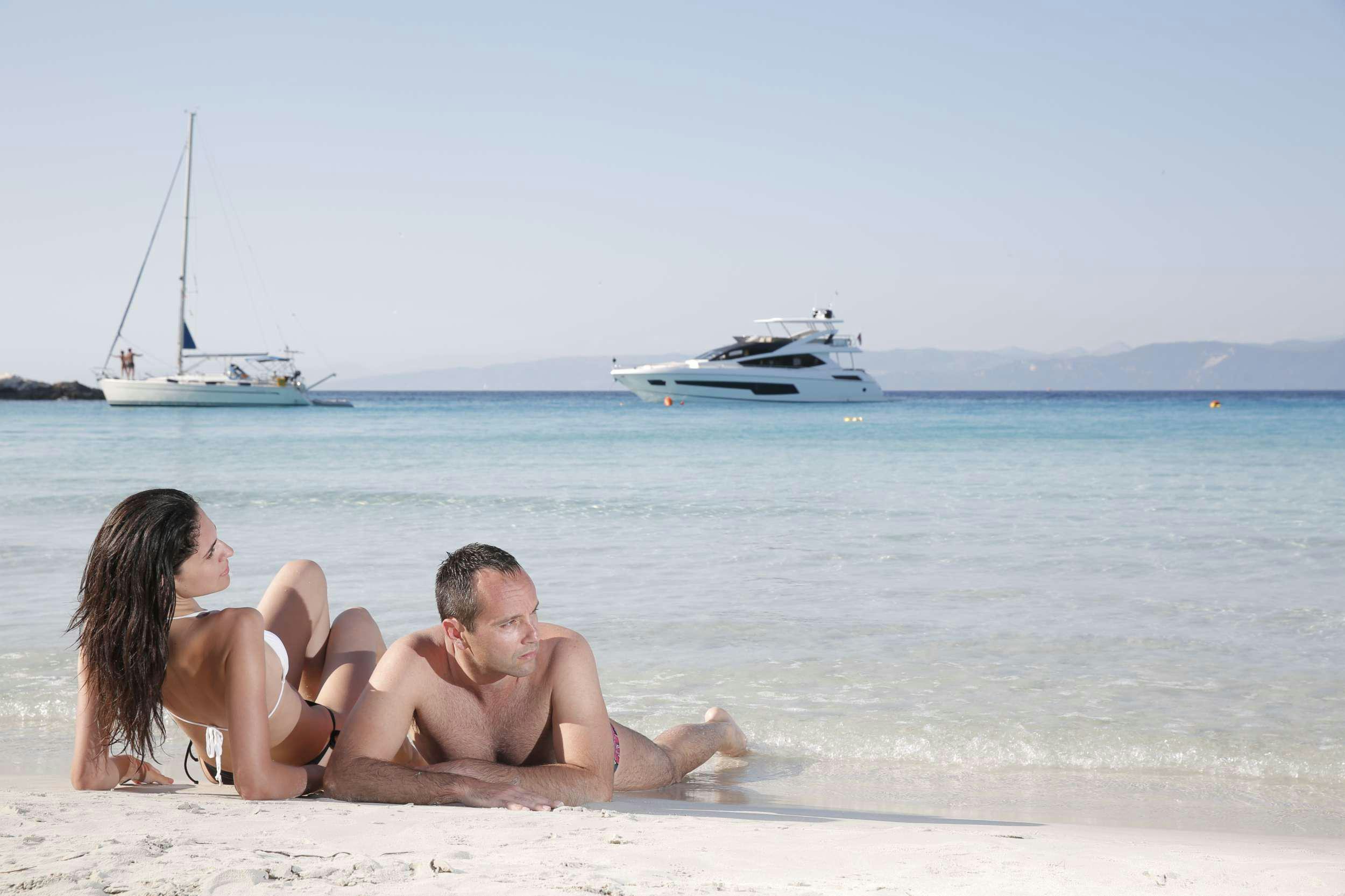 a man and woman sitting on a beach with a boat in the background aboard FINEZZA Yacht for Charter