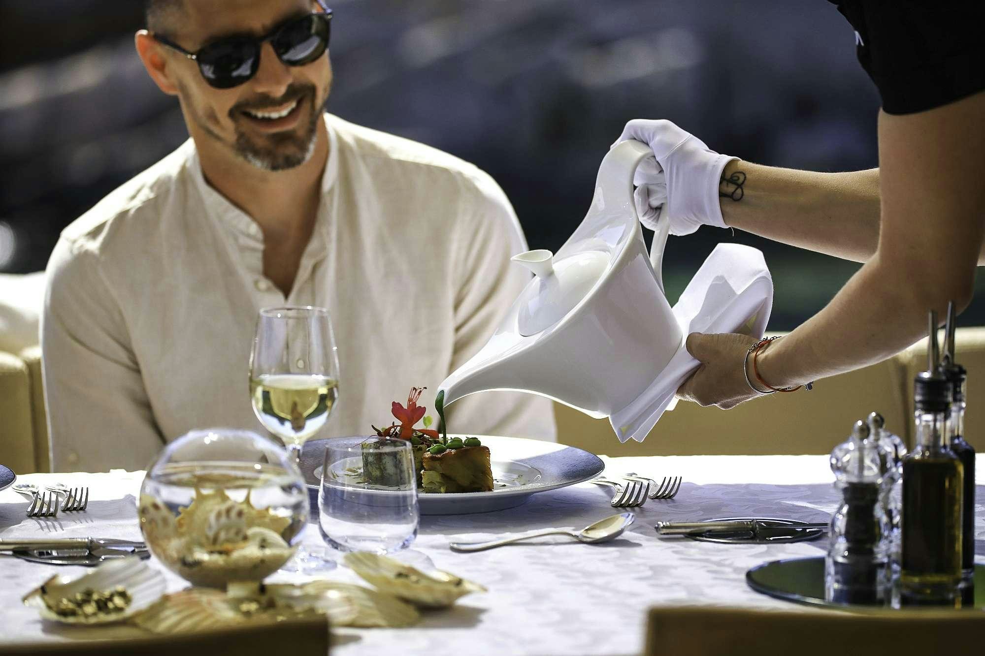a person in a white robe sitting at a table with food and a bottle of wine aboard AGAVE Yacht for Charter