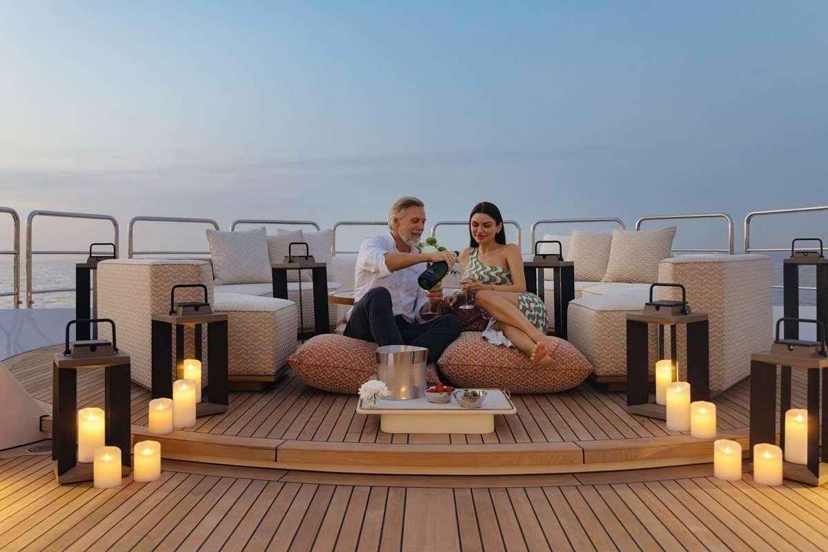 a man and woman sitting on a couch on a balcony with a beach view aboard BOMBAY Yacht for Charter